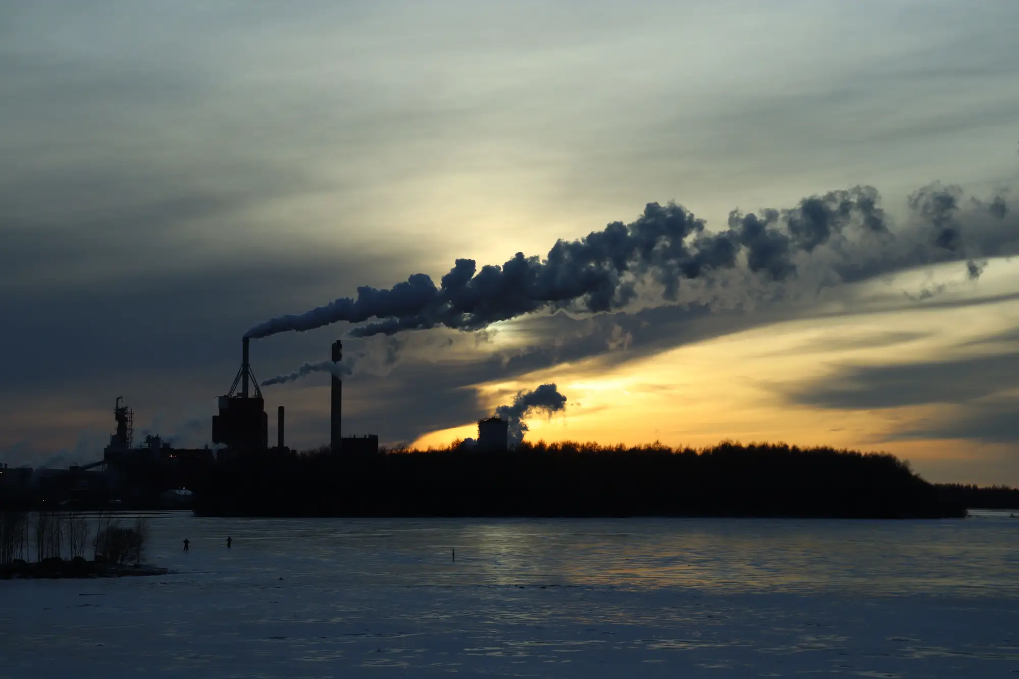 Silhouetted industrial plant on a frozen shoreline, chimneys trailing plumes into a winter sunset